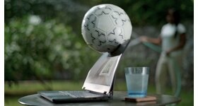 Picture of a Dell laptop over a table with a glass cup behind the product. A soccer ball is hitting the laptop screen.