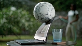 Dell laptop over a table with a glass cup behind the product. A soccer ball is hitting the laptop screen.
