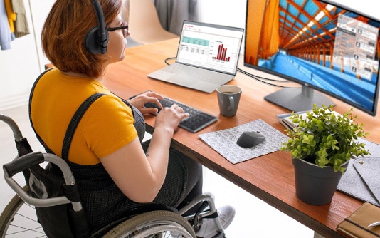 Kvinde i kørestol ved sit skrivebord, der arbejder på en bærbar Dell-computer og skærm.