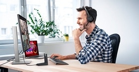 Smiley man with a headset on his head using a Dell monitor, mouse and keyboard placed over a wood table.