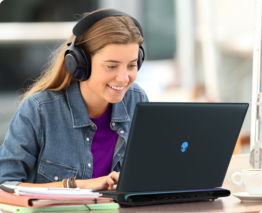 Happy student wearing headphones using an Alienware laptop at a desk with notebooks and a coffee cup, listening a video tutorial.