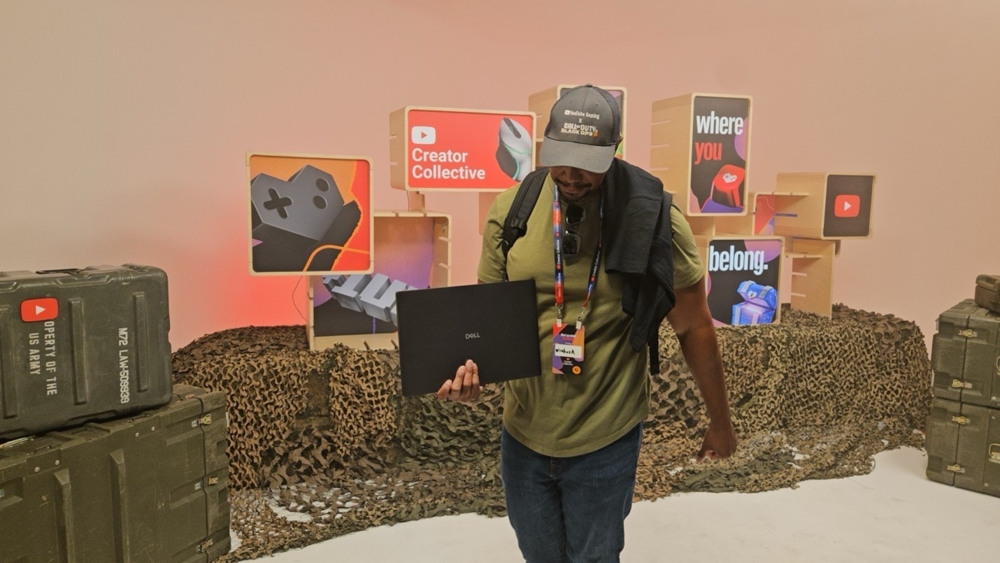 A man holds a Dell laptop at a YouTube Creator Collective event with military crates and camouflage.