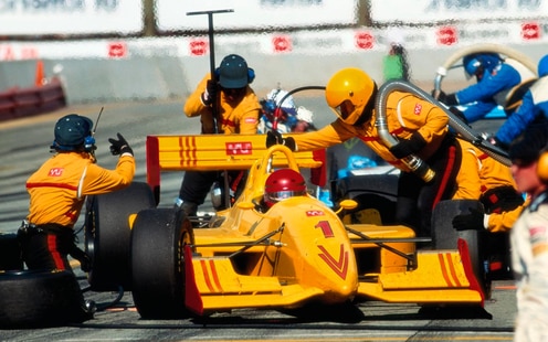 F1 race car team in yellow outfits work on the yellow race car with red stylized design during a pit stop.
