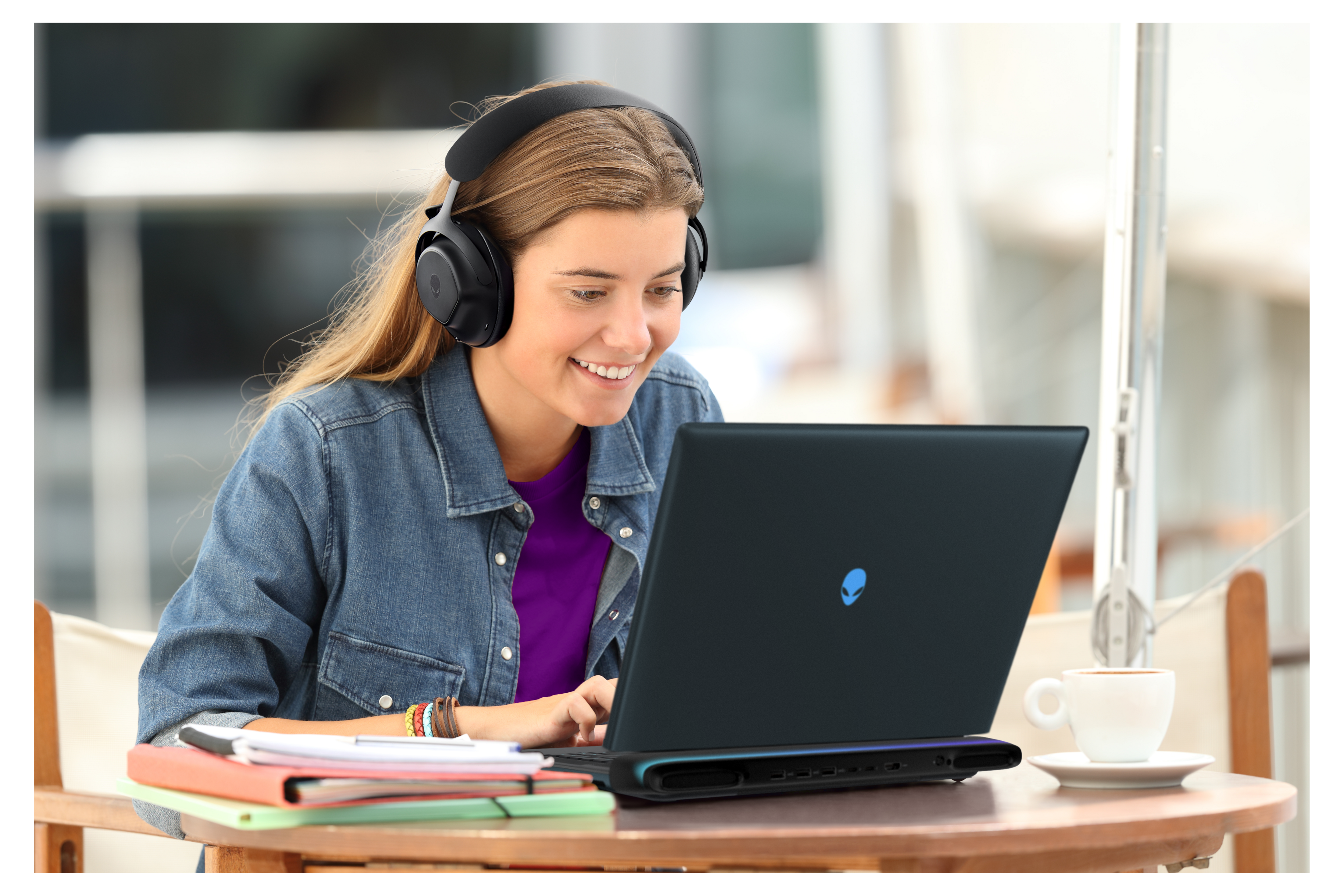 Happy student wearing headphones using an Alienware laptop at a desk with notebooks and a coffee cup, listening a video tutorial.