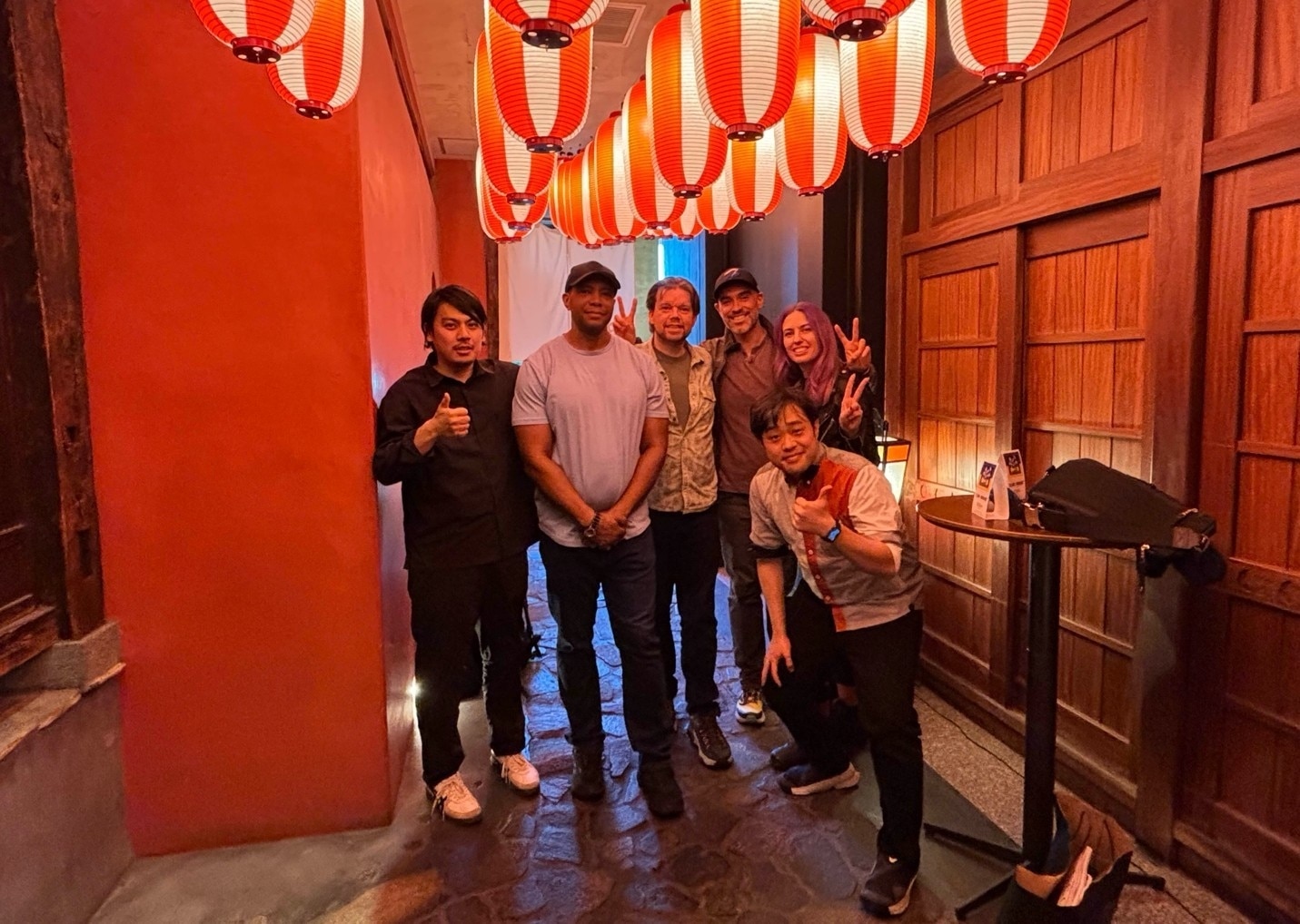 Six people pose for a photo in a cobblestone hallway illuminated by hanging red paper lanterns.