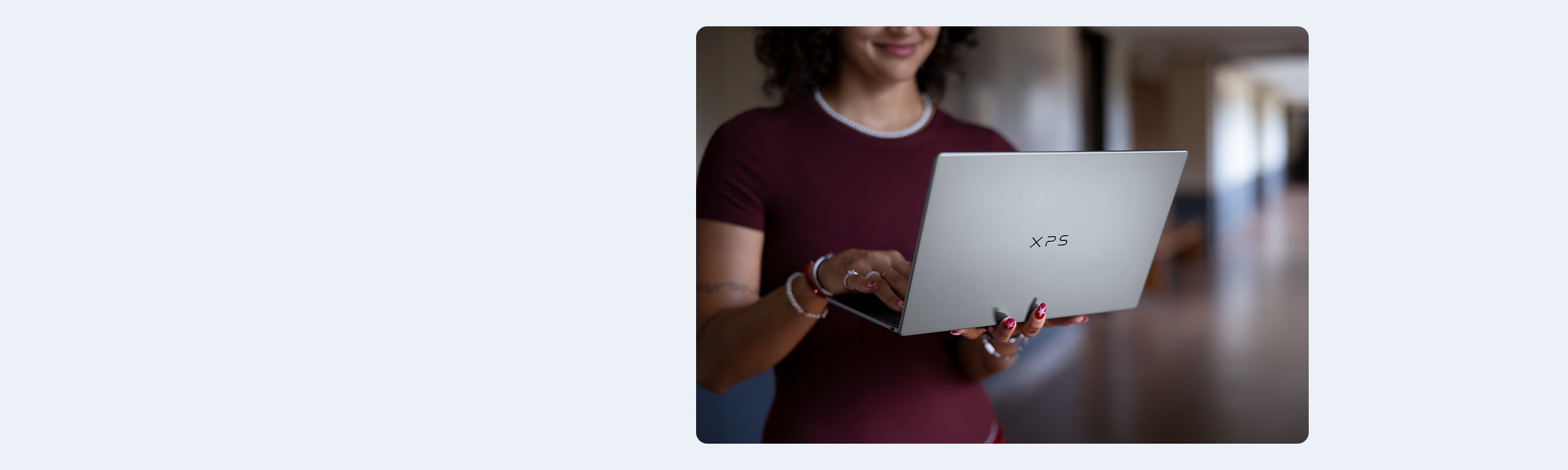Smiling woman in a maroon shirt holding and typing on a silver Dell XPS laptop in a hallway.
