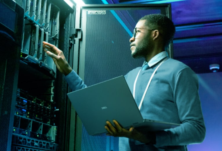 Man in a server room holds a silver Dell laptop while checking a server rack illuminated by blue and green lights.