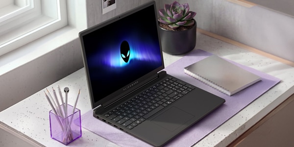 Open Alienware laptop on a white desk with a notebook, succulent, and pen holder beside window. 
