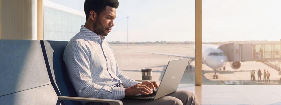 A man works on a silver Dell laptop in an airport terminal with an airplane visible through the window.