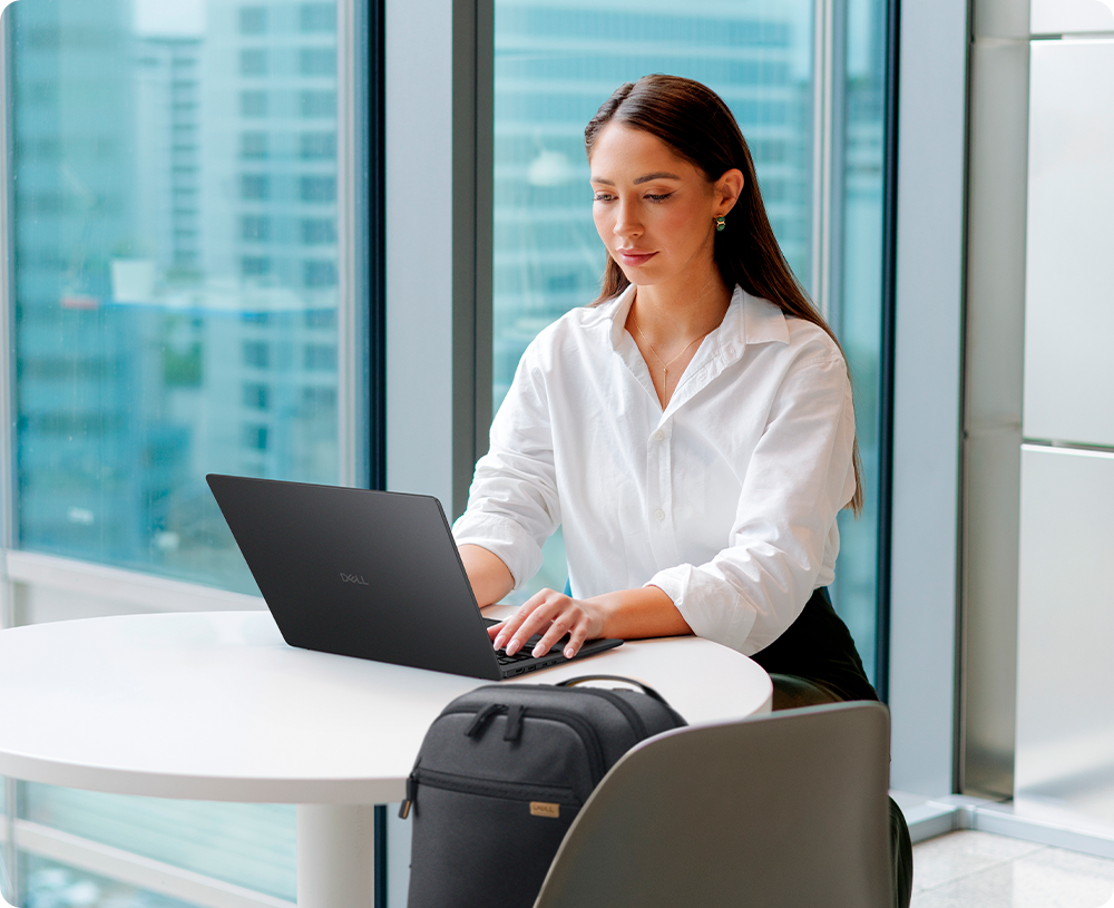 Woman using Dell Pro P516265 Laptop at a white table in a modern office with large windows.