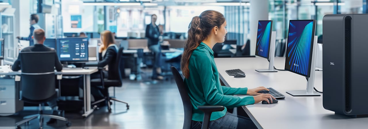 Woman using Dell desktop in a contemporary office with dual monitors and coworkers in background.