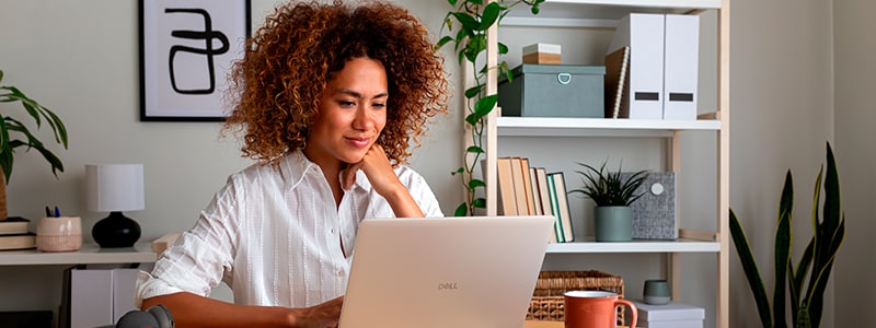 Woman working at a Dell laptop in a home office with shelving, books, and plants.