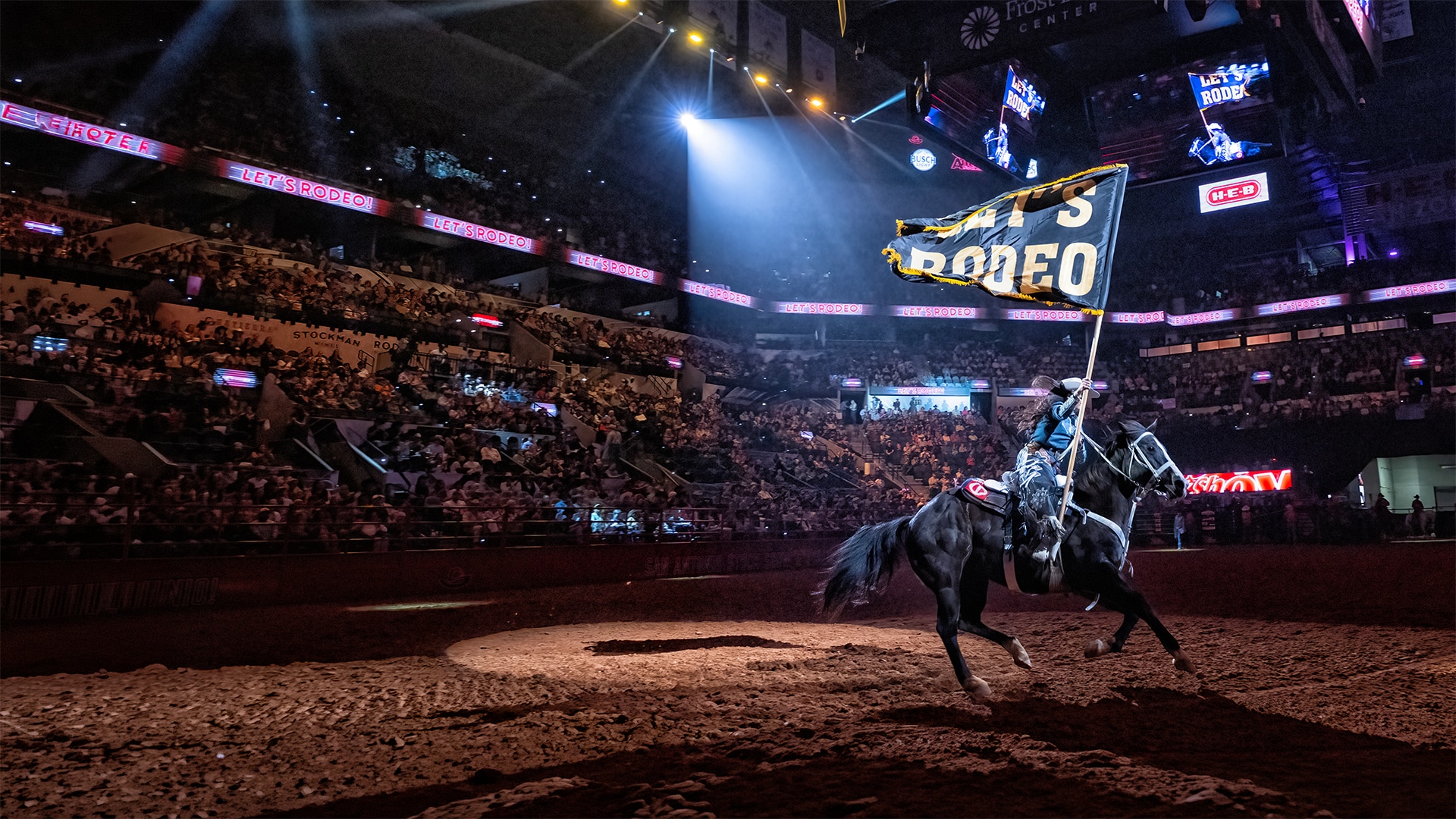 A rider on a black horse waves a flag in a bright rodeo arena packed with spectators.​