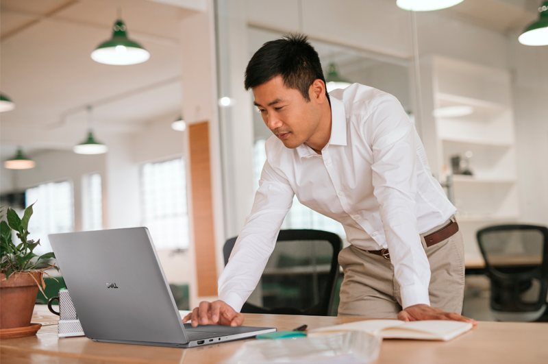 Man in a white shirt standing at a desk in a bright office, working on a silver Dell laptop under green pendant lights.