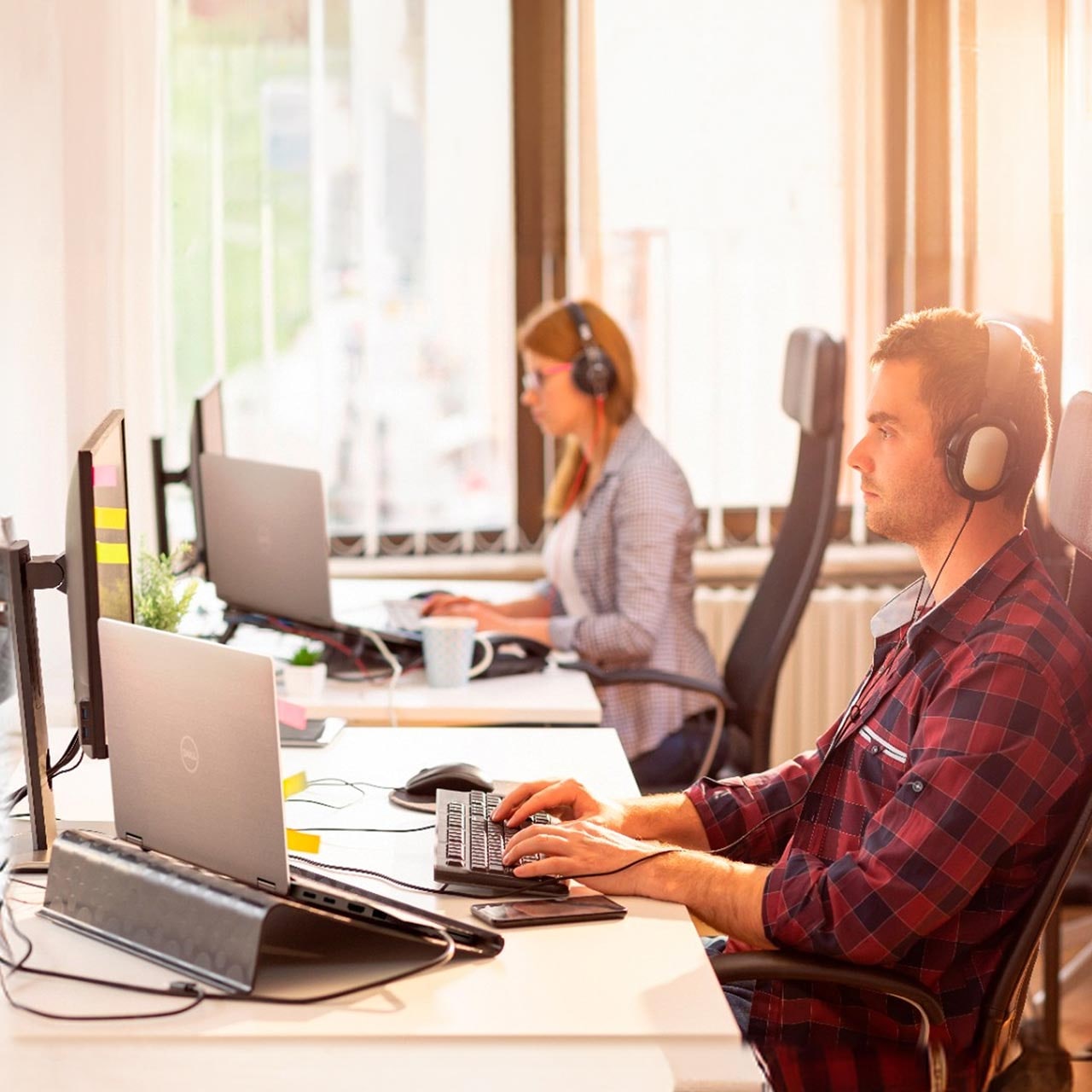 Man in a headset and plaid shirt types on a keyboard at a desk with a Dell laptop, with a coworker in the background.