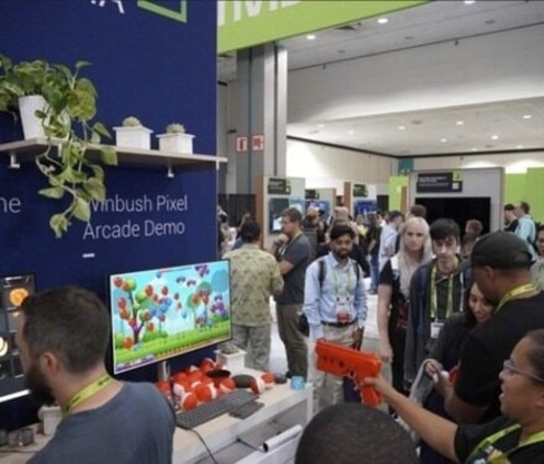 A person points an orange light gun at a computer monitor displaying a balloon-popping game at a crowded event booth.