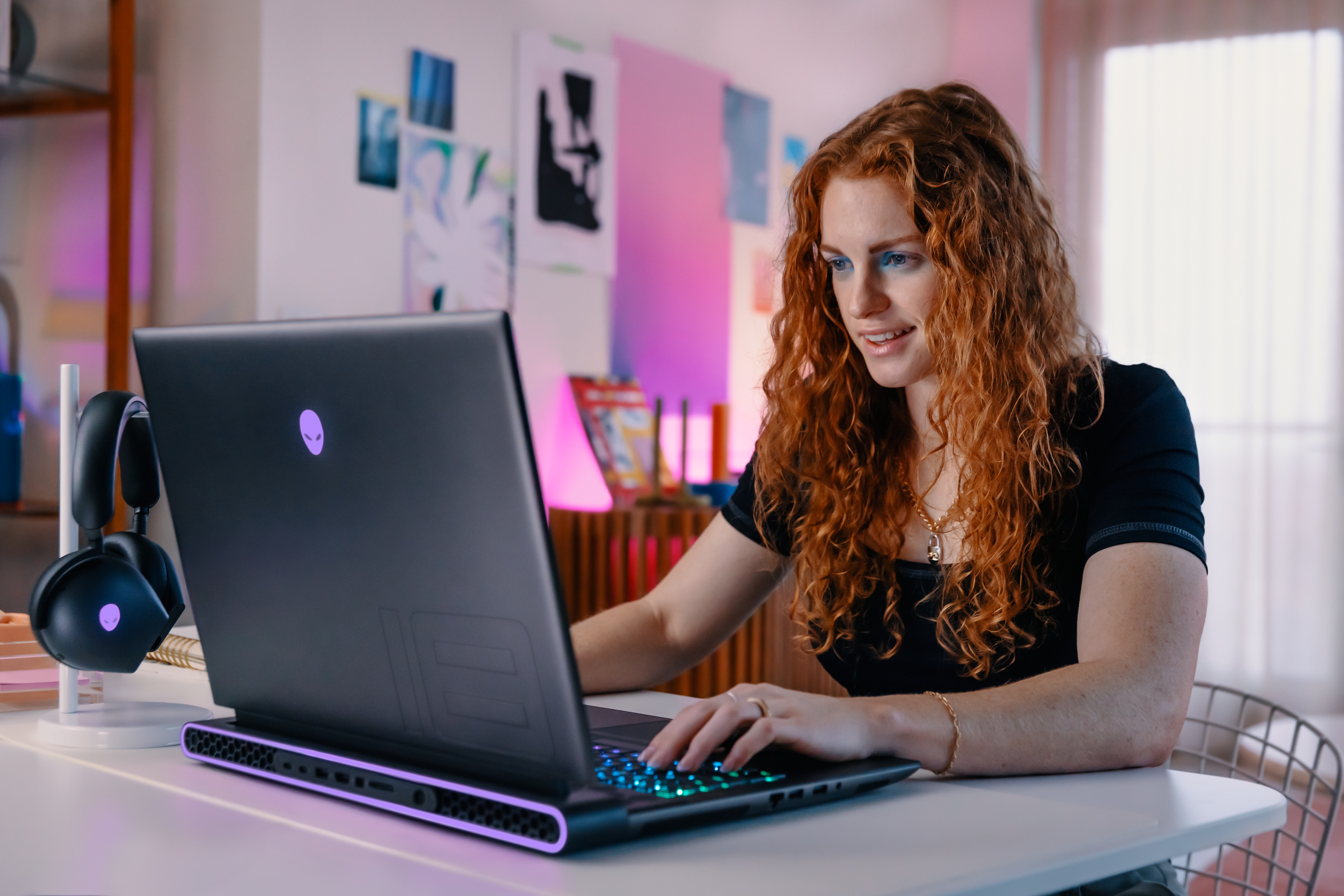 Woman in a bright room using an Alienware gaming laptop with headphones hanging next to her.