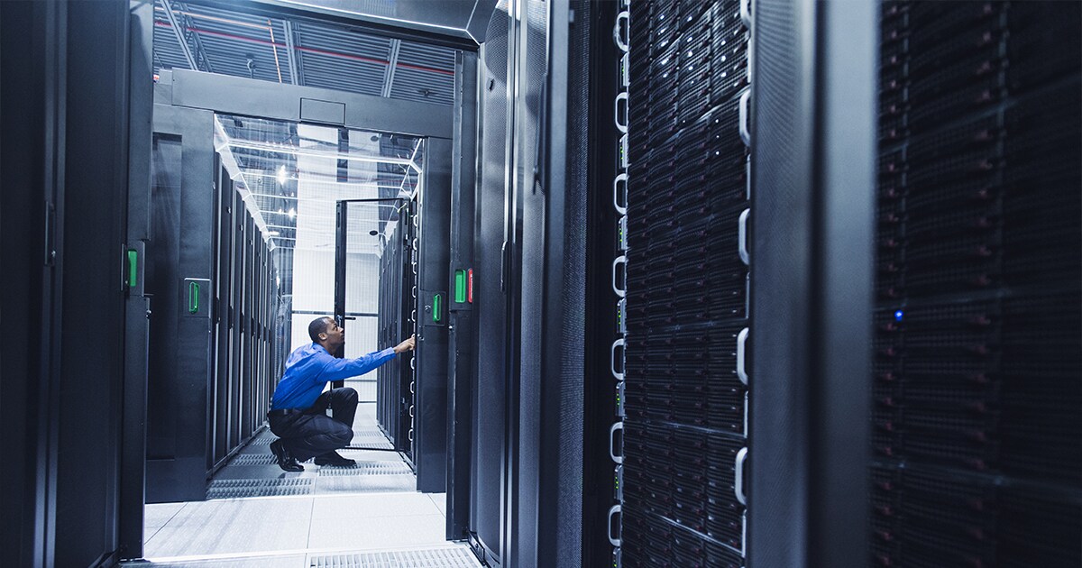 Image of a man in a blue button-down shirt in a room of servers pushing buttons on one of the servers. 