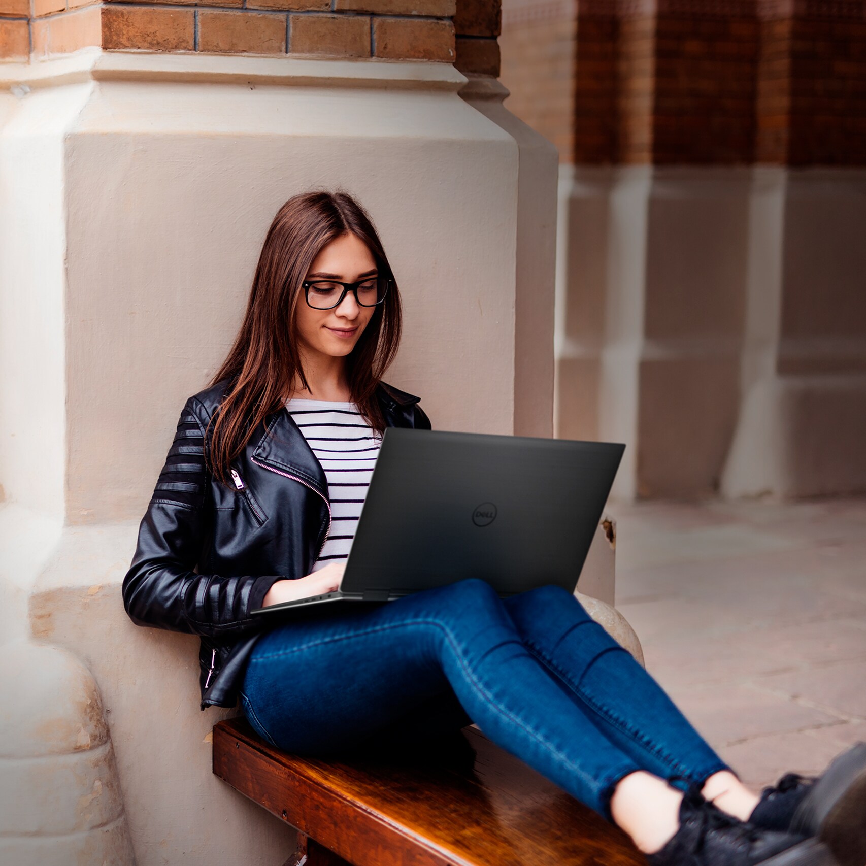 A woman working on her laptop while sitting on a bench outside of a university building.
