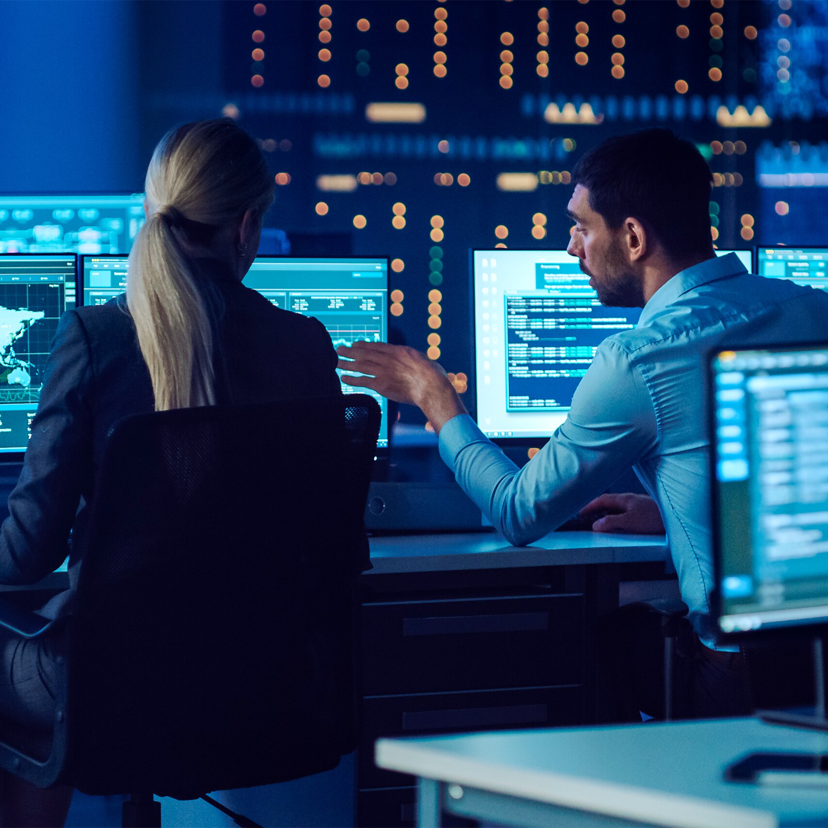 Two people in a control room analyze data on multiple computer screens, with a backdrop of illuminated control panels and monitors.