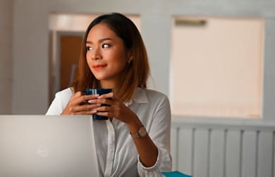 Woman with a cup in her hands using a Dell laptop on a table in front of her.