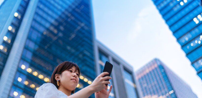 A Young woman is using a smart phone in front of tall skyscrapers.