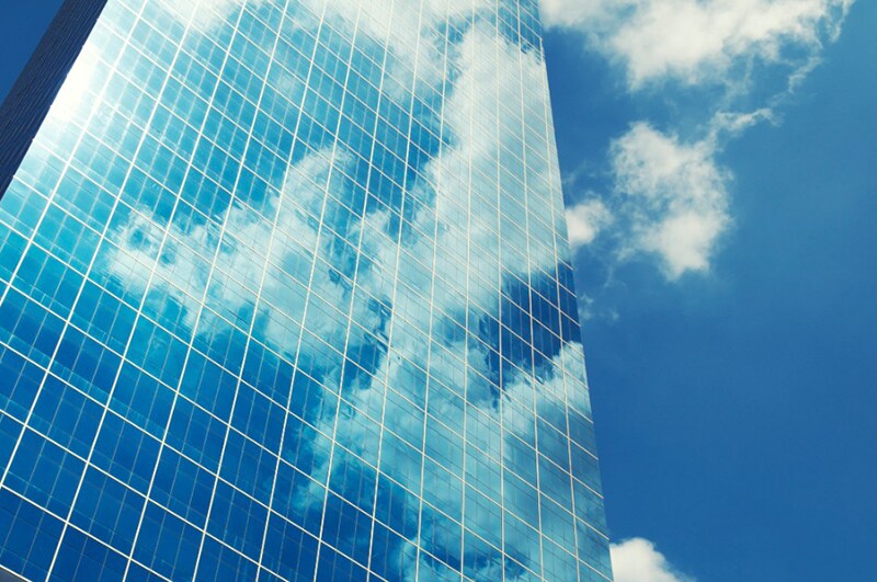 Sleek modern glass and steel office skyscraper jutting into blue sky reflecting fluffy white clouds making a business reflection