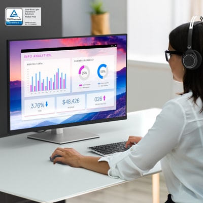 Picture of a woman using Dell products over a table, including a Dell P3223QE monitor and a Dell keyboard and mouse.