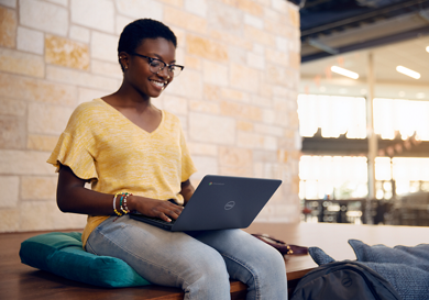 Picture of a seated woman wearing a yellow t-shirt and jeans smiling with a Dell laptop on her lap. Behind her, a brick wall.