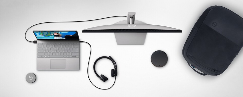 Imagem de um notebook Dell com headset e monitor conectados, uma mochila, um adaptador móvel e um alto-falante — vistos de cima.