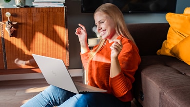Picture of a smiling woman wearing an orange sweater sitting on the floor with a Dell Inspiron 14 5420 Laptop on her lap.