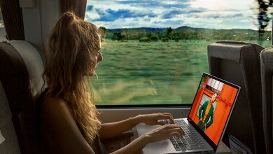 Picture of a woman on a train using a Dell laptop on the coffee table in front of her. A large window shows the landscape.