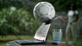 Picture of a Dell laptop over a table with a glass cup behind the product. A soccer ball is hitting the laptop screen.