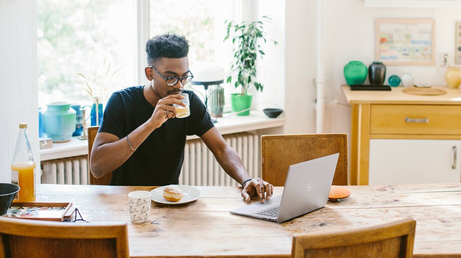 Imagen de un hombre con un vaso de jugo en la mano derecha con una laptop Dell Inspiron 16 5620 en una mesa frente a él.
