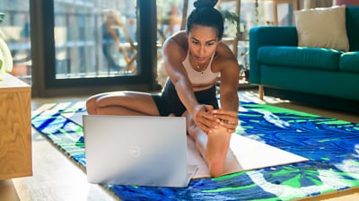 Picture of a woman in a living room stretching herself with an Inspiron 16 5620 Laptop on the carpet in front of her.