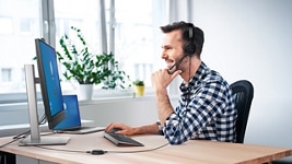 Picture of a smiley man with a headset on his head using a Dell monitor, mouse and keyboard placed over a wood table.