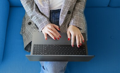 Zdjęcie kobiety, która siedzi na niebieskiej kanapie z notebookiem Dell Vostro 16 5620 na kolanach i pisze na jego klawiaturze.