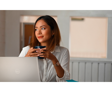 Kobieta trzymająca w ręku kubek korzysta z notebooka Dell stojącego przed nią na stole.