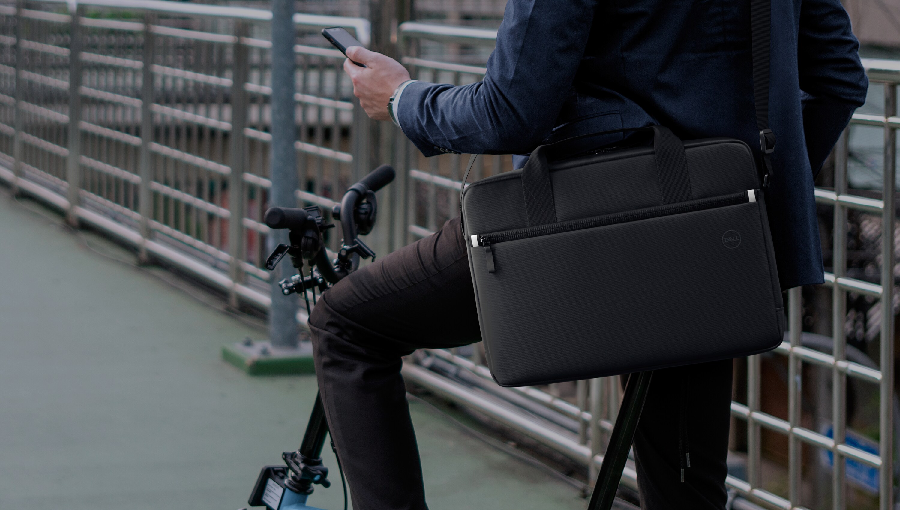 Man on a bicycle holding a cell phone in his left hand with a briefcase across his chest.