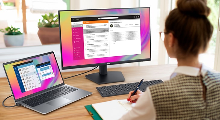 Picture of a woman using a Dell SE2723DS Monitor connected to a Dell Laptop on a wood table. 