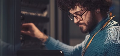 Sol elinde bir tablet ve sağ eli duvardaki kablolarda olan mavi süveterli ve gözlüklü bir adamın resmi.