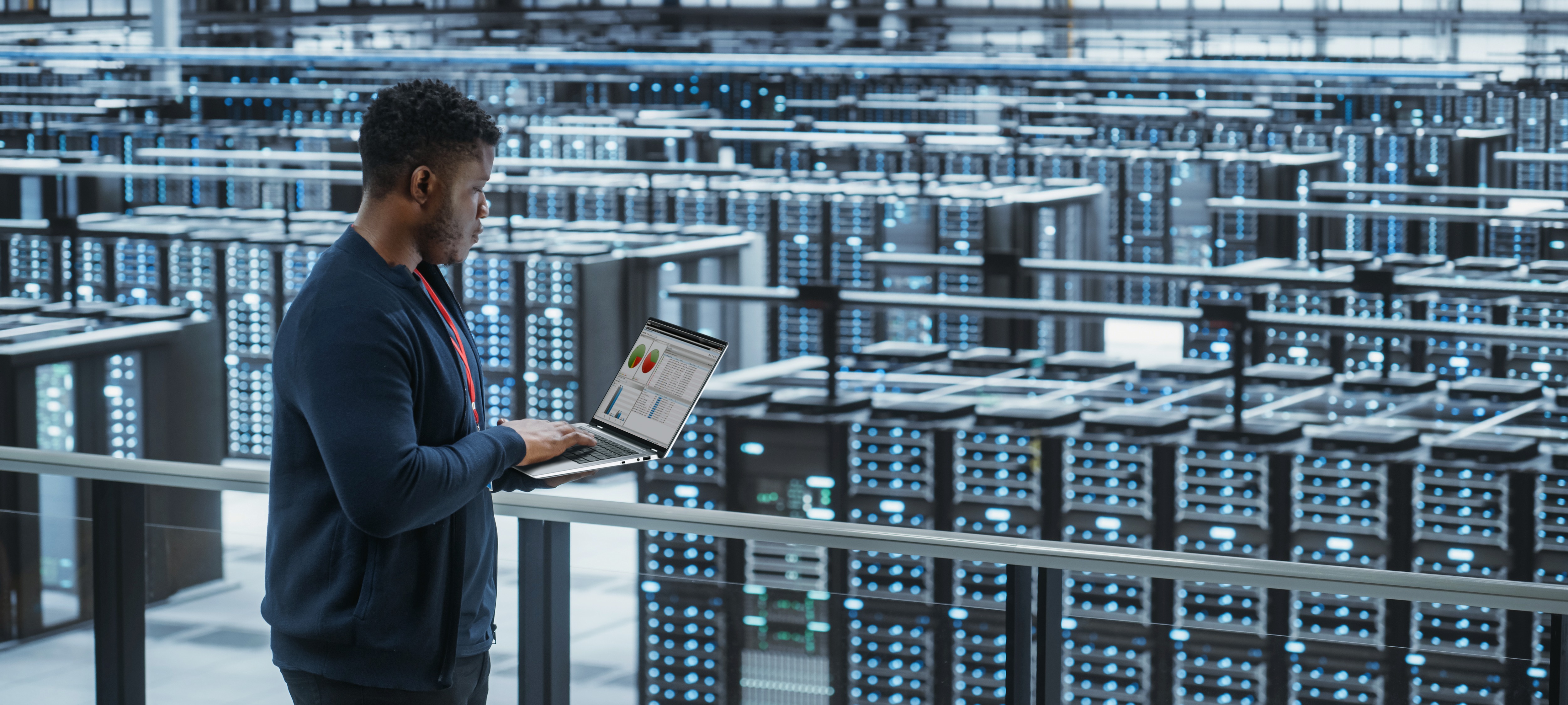 Homem em uma sala de servidores segurando um laptop e digitando nele.