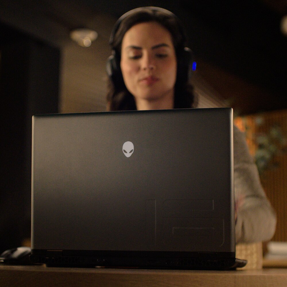Low-angle, rear side view of an Alienware M16 R2 laptop on a table within a dimly lit room, a woman wearing a headset is sat using it. 