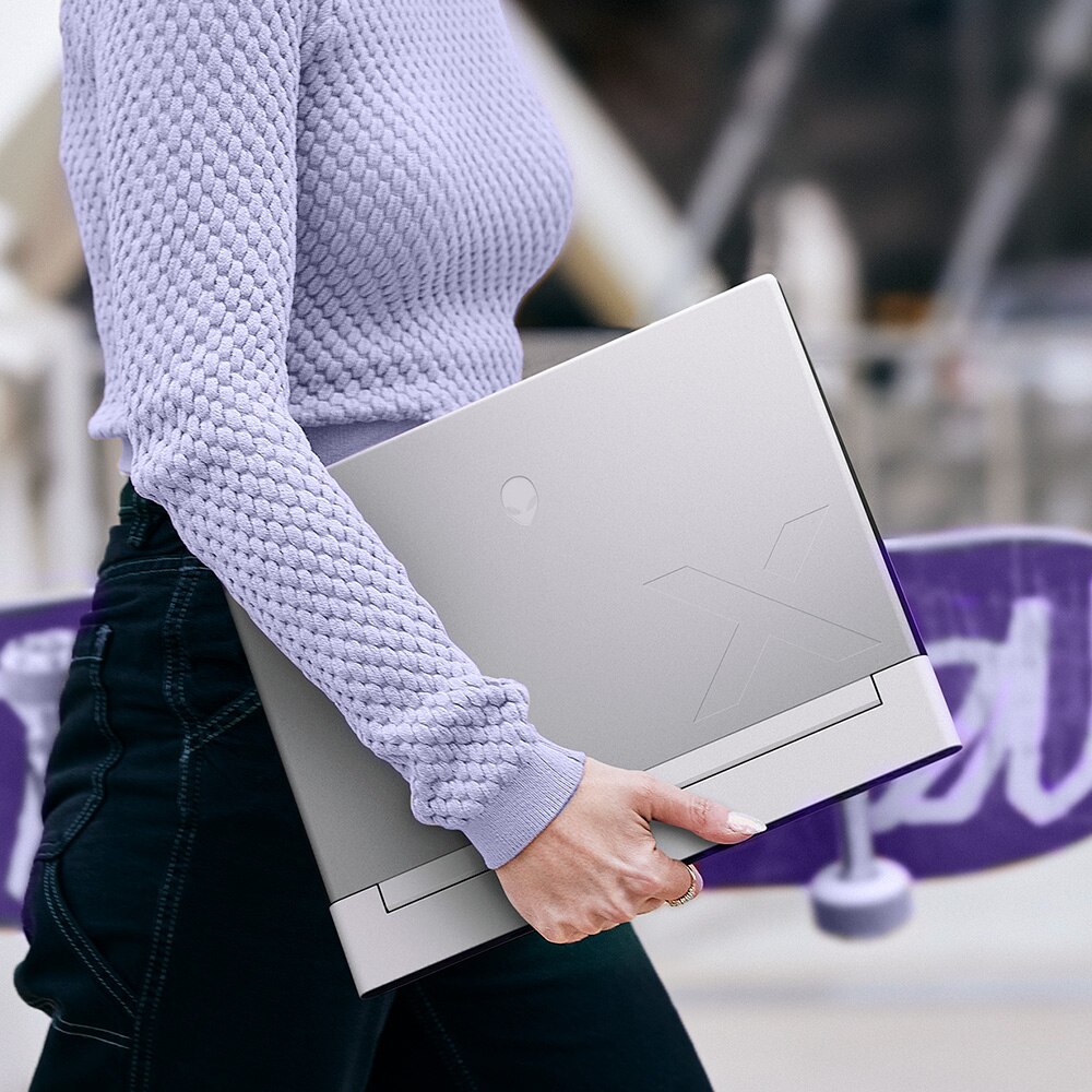 A woman, only her mid-section in frame, carries an Alienware X14 R2 laptop in one hand and a skateboard in the other as she walks through an urban setting.