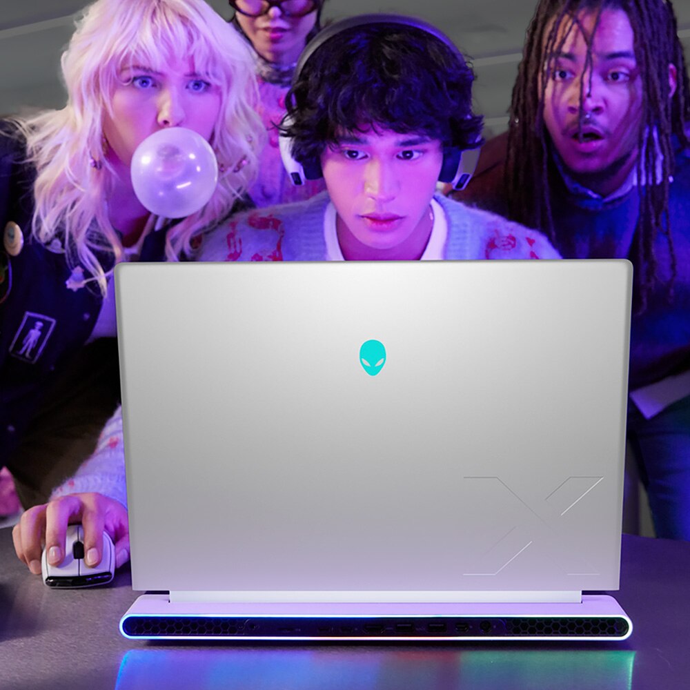 Rear side view of an Alienware X16 R2 laptop, a young man plays a game intently as three friends watch with anticipation from behind.