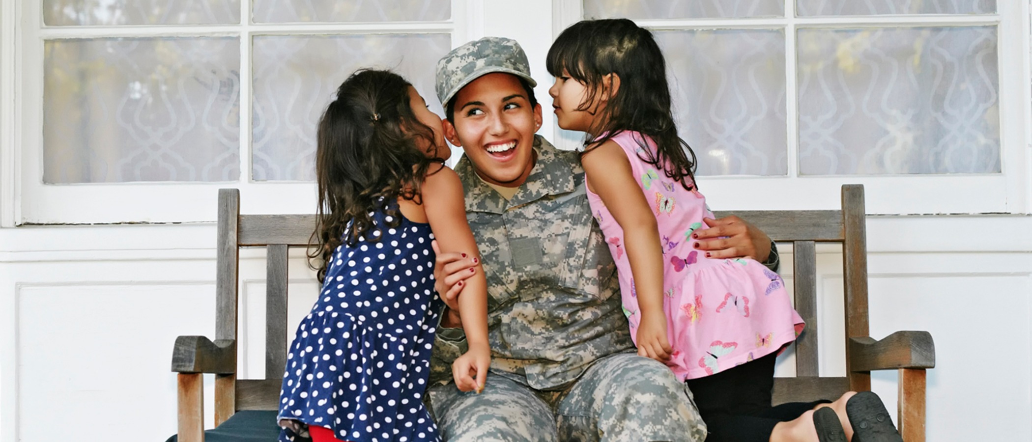 Smiling woman in military camouflage uniform seated as two young girls lean in to kiss her on the cheeks.