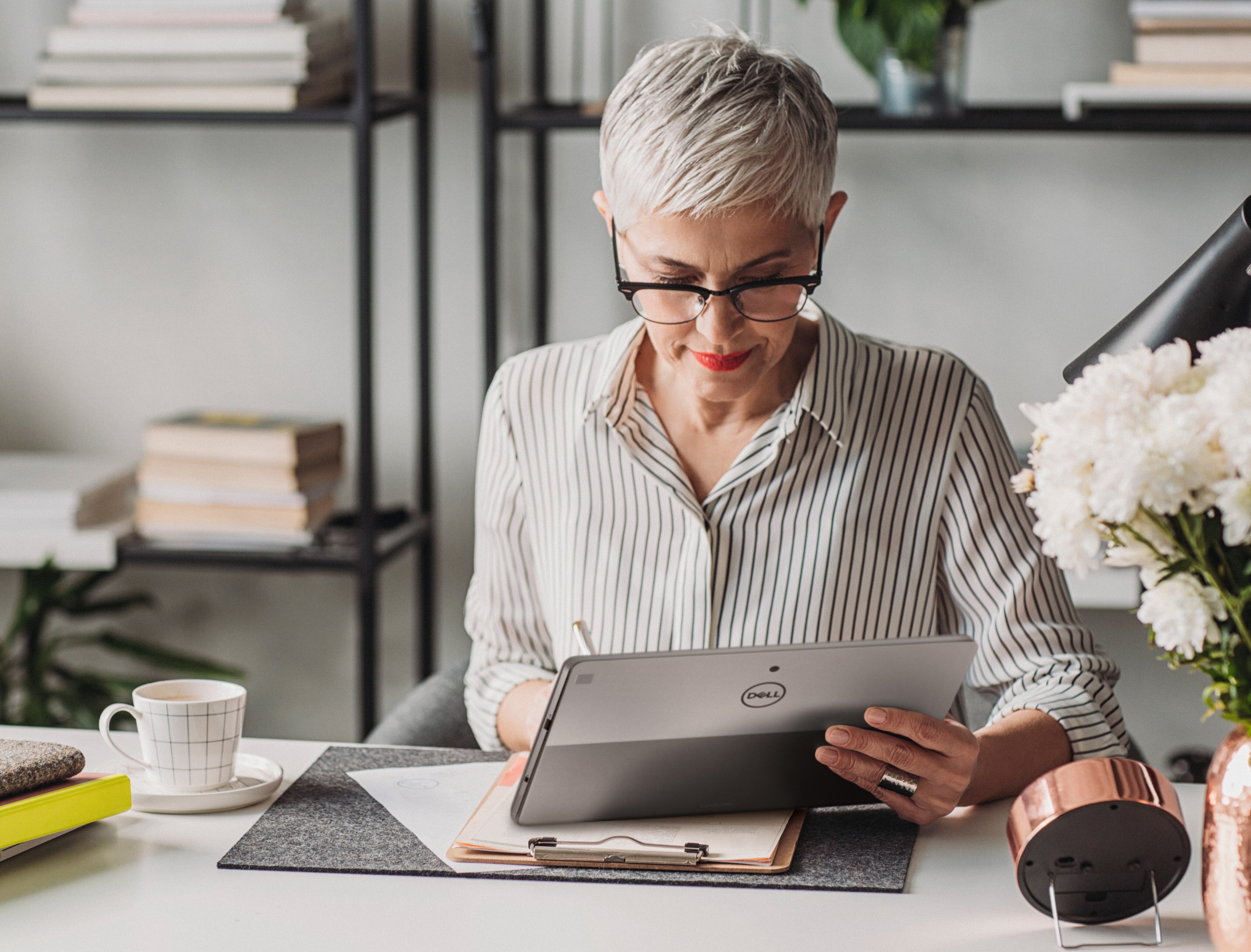 Picture of a sitting woman at the office with a Dell Tablet on her left hand and writing on a paper with the right hand. 