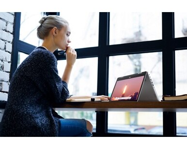 Afbeelding van een zittende vrouw met een Dell 2-in-1 laptop opengeklapt als een tablet boven een houten tafel voor haar.