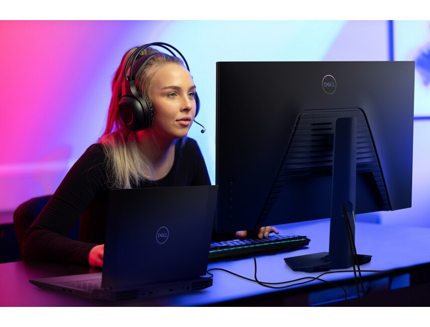 Picture of a sitting woman with a headset on her head and a Dell Monitor, a Dell Laptop, Keyboard, and mouse on a table.