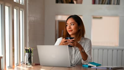 Imagem de uma mulher com um copo nas mãos usando um notebook Dell XPS 13 9320 em uma mesa à frente dela.